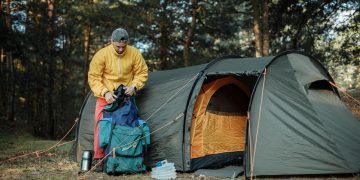 A young man organizing camping gear beside a tent in a scenic forest.