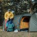 A young man organizing camping gear beside a tent in a scenic forest.